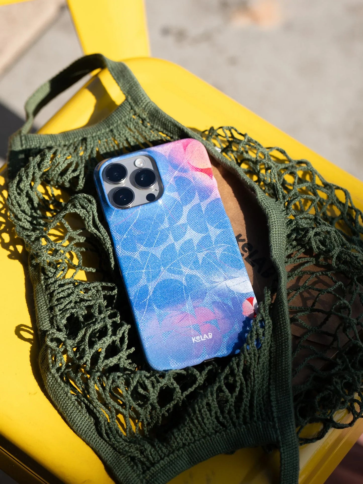 KELAB retro blue iPhone case with abstract poolside design resting in a green mesh bag, showcasing summer-ready accessory style.