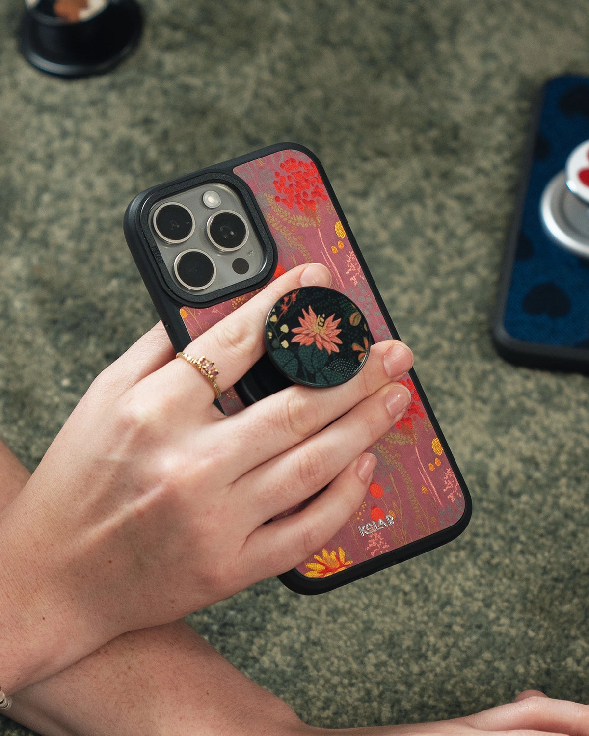 Hand holding a phone with a KELAB phone case that features flowers on a pink backdrop and a pop socket