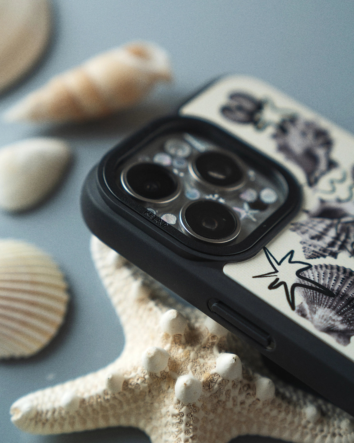 KELAB iPhone case with seashell design on a gray surface with shells and a starfish.