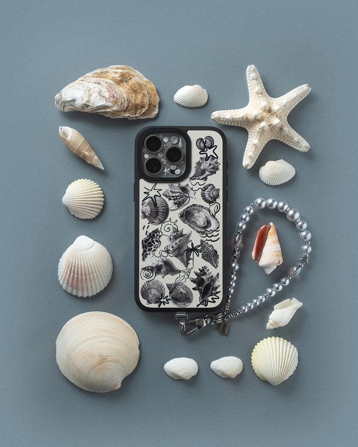 KELAB brand iPhone case with sea-themed design surrounded by seashells and a starfish on a gray background